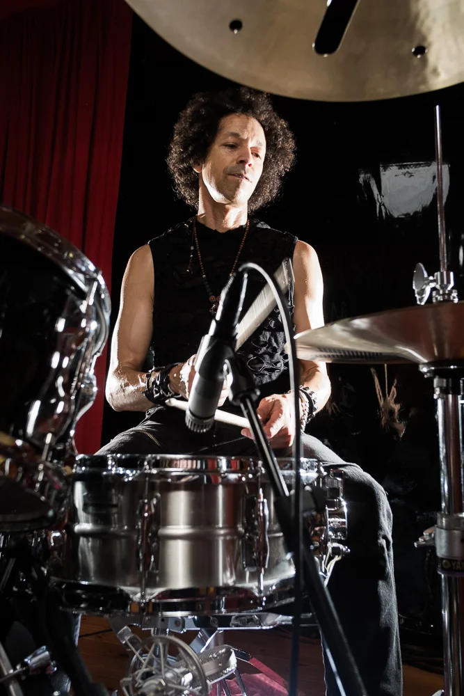 Zack playing drums. The shot points up from a low angle through drum kit, framing Zach between cymbals and bass drum.